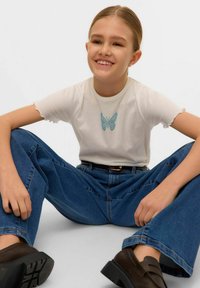 T-shirt blanc avec des manches à volants, orné d'une broderie de papillon bleu. Porté avec un jean bleu taille haute et des chaussures marron foncé.