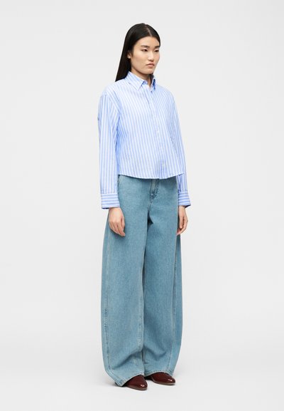 Γυναίκα που φοράει μπλε και λευκό πουκάμισο με κάθετες ρίγες, φαρδιά ανοιχτόμπλε τζιν παντελόνια και σκούρα καφέ παπούτσια, στέκεται σε απλό φόντο.