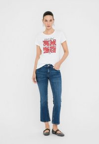 T-shirt bianca con stampe grafiche in rosso e nero, abbinata a jeans in denim blu con taglio dritto e orlo sfilacciato. Ballerine nere.