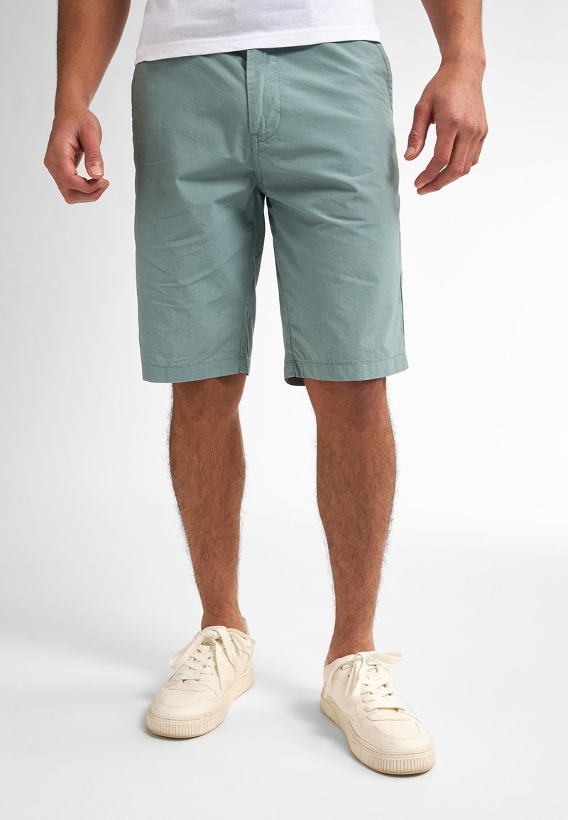 Shorts en coton vert clair avec poches avant, fermeture à bouton et coupe décontractée ; associés à des baskets blanches et un t-shirt blanc uni.