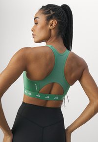 Soutien-gorge de sport athlétique vert avec un design dos nageur, détail découpé et une bande à motifs avec des accents réfléchissants. Des leggings noirs sont également visibles.