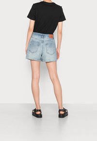 Light blue denim shorts with back pockets, slightly frayed hem, and a brown leather brand patch at the waistband. Black sandals are worn.
