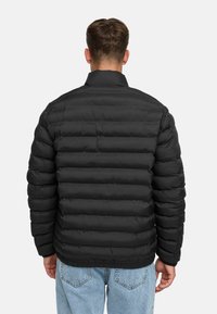 Sort pufferjakke med vandret quilting, ståkrave, ribbede manchetter og elastisk kant. Båret over lyseblå jeans.