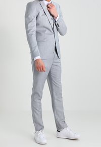 Světle šedý oblek s slim fit, texturovaná tkanina, jednopřeska sako a odpovídající kalhoty, doplněné bílými teniskami.