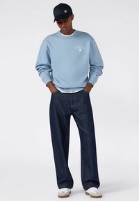 Felpa color azzurro chiaro con logo, vestibilità comoda, abbinata a jeans scuri in denim e sneakers bianche, accessoriata con un cappello nero.