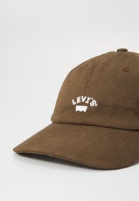 Brun cap laget av teksturert stoff med en buet visir. Har hvit brodert "LEVI'S" logo foran. Klassisk baseballhattdesign.