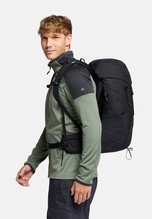 Sac à dos noir avec plusieurs poches zippées. Porté sur une chemise à manches longues verte avec des accents noirs. Comprend des bretelles réglables et un dos en mesh.