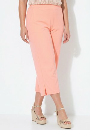 Vrouw draagt lichtroze korte broek met vooraan splits en beige sleehakken tegen een witte achtergrond.