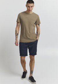 T-shirt en coton brun avec un col rond et une poche poitrine, associé à des shorts roulés navy et des baskets noires. Tatouages visibles sur les bras.