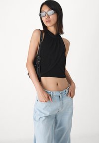 Black sleeveless top with a high neckline and draped design, paired with light blue loose-fit jeans. Model holds pockets, wearing sleek eyewear.