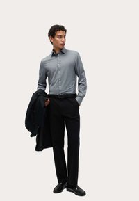 Grijze langemouwen shirt met knoopsluiting, zwarte broek en glanzende zwarte schoenen. Persoon houdt een zwart jasje vast en staat in een ontspannen houding.