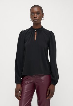 Schwarze Bluse mit langen Ärmeln, Volantkragen, Ausschnitt mit Schlitz und elastischen Bündchen, kombiniert mit burgunderfarbenen Kunstlederhosen.