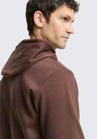 Bruine hoodie met een ontspannen pasvorm, voorzien van een trekkoordkap, een gladde textuur en gestikte naden, gezien vanuit de zijkant en de achterkant.