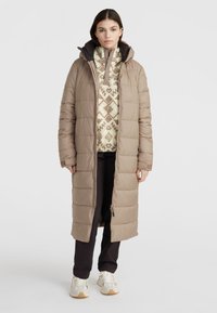 Lång beige puffjacka med huva, med quiltat mönster, dragkedja fram och ståkrage, bärs över en mönstrad topp och svarta byxor.