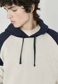Haut à capuche beige et marine avec un cordon de serrage, présentant un corps crème et des manches navy. Comprend un petit logo discret à l'avant.