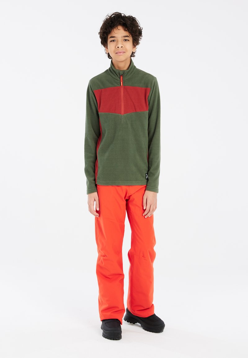 Sudadera de forro polar verde con panel rojo en el pecho, cuello con media cremallera; combinada con pantalones naranjas brillantes y botas de invierno negras. Textura suave, ajuste casual.