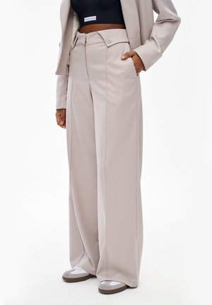 Osoba ubrana w beżowe spodnie o szerokich nogawkach z zapinanym na guziki pasem, czarny krótki top, rękaw beżowej marynarki i białe szaro-białe sneakersy, ręka w kieszeni.