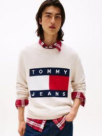 Kreemjas kootud sviiter, millel on merevaigukollane ja punane TOMMY JEANS logo, kiht kaitseb punase ruudulise särk üle. Näha on tume sinine teksapüks.