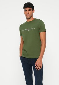 Olivově zelené bavlněné tričko s krátkým rukávem, s bílým textem a logem "TOMMY HILFIGER". Klasický design s kruhovým výstřihem. Nosené modré džíny.