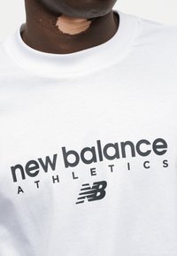 Λευκό βαμβακερό μπλουζάκι με μαύρο κείμενο "new balance ATHLETICS" και λογότυπο, στρογγυλή λαιμόκοψη, ομαλή υφή και κοντά μανίκια.
