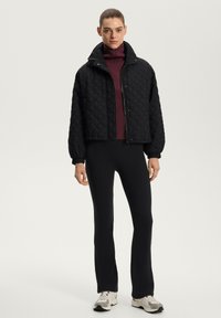 Jachetă scurtă, cu model matlasat, de culoare neagră, cu guler înalt, purtată cu un pulover bordeaux cu guler înalt și pantaloni negri ajustați. Material texturat, croi lejer.