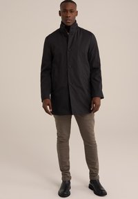 Abrigo negro impermeable con un alto cuello, botones frontales y bolsillos laterales; combinado con pantalones marrones claros y botas negras. Textura suave.