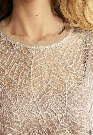 Haut beige avec une superposition texturée et transparente arborant un motif de feuilles et un léger éclat. Encolure ronde et bords délicats complètent le design.