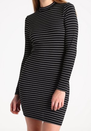 Femme portant une robe noire moulante à manches longues et côtelée avec des rayures horizontales blanches, debout devant un fond blanc uni.