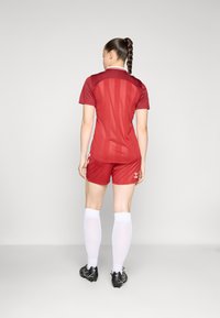 Camisola atlética vermelha de mangas curtas e colarinho, combinada com calções vermelhos. Meias brancas até aos joelhos e botas pretas completam o conjunto.