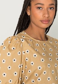 Blusa bege com mangas bufantes, adornada com flores brancas e centros pretos. Textura lisa com decote redondo.