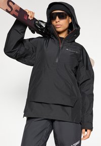 Schwarze wasserdichte Jacke mit einem Reißverschluss vorne und verstellbarem Kapuzen, ausgestattet mit einer lockeren Passform und einem auffälligen Logo auf der Brust, kombiniert mit Skiausrüstung.