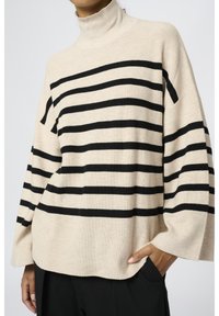 Beżowy sweter z golfem w czarne paski, posiada prążkowaną fakturę, szerokie rękawy oraz boczne kieszenie dla praktycznego zastosowania.