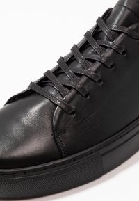 Gros plan d'une basket en cuir noir avec des lacets noirs texturés et une semelle épaisse en caoutchouc sur fond blanc.