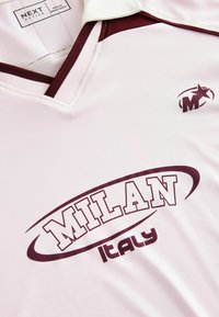 Světle růžový sportovní dres s výstřihem do V, vínovými akcenty, na kterém je v tučném písmu vytištěno "MILAN" a "Itálie". Logo se nachází v levém horním rohu.