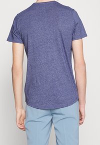 Lila T-Shirt mit kurzen Ärmeln aus leichtem Stoff, mit rundem Ausschnitt und abgerundetem Saum, von hinten betrachtet.