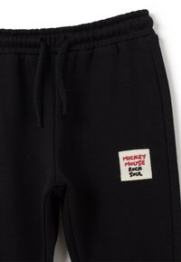 Pantalons de survêtement noirs en coton, avec une patch de marque portant "MICKEY MOUSE" et "Rock Soul" en rouge sur un fond beige. Ceinture élastique.