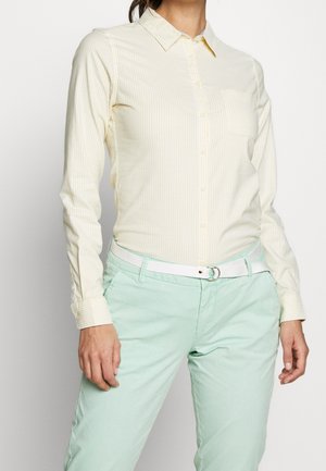 Kvinde iført en lys gul, stribet skjorte med knapper, stoppet ned i mintgrønne bukser med et hvidt bælte, stående mod en ensfarvet baggrund.