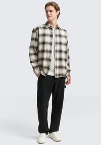 Beige und schwarze karierte Flanellhemd mit Knopfverschluss, über einem weißen T-Shirt getragen; kombiniert mit schwarzen Hosen und weißen Sneakers.