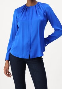 Kvinna som bär en ljusblå långärmad blus med samlad halsringning och mörka slim-fit jeans, poserande med en hand på höften.