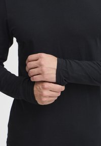 Bluza neagră cu mânecă lungă, confecționată dintr-un material ușor, cu manșete ajustate și o textură netedă, având mâinile poziționate aproape de torace.