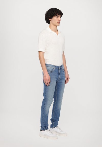 Polo shirt à manches courtes, de couleur beige clair, avec une texture douce, associé à un jean slim-fit bleu moyen et des baskets blanches.