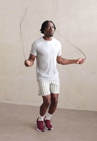 En atletisk mand hopper i reb iført en grå Nike t-shirt, stribede shorts og kastanjefarvede sneakers med hvide sokker foran en neutral baggrund.