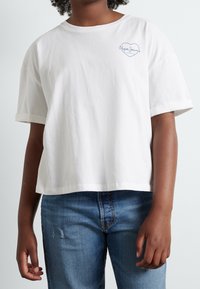 Valkoinen puuvillainen t-paita, jossa on käännetyt lyhyet hihat, sininen sydänlogo vasemmassa rinnassa ja rento istuvuus. Päällä siniset farkut.