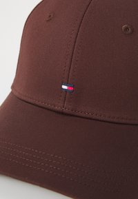 Brown baseball cap with a curved brim, featuring a small embroidered logo in red, white, and blue. Made of textured fabric with stitching details.