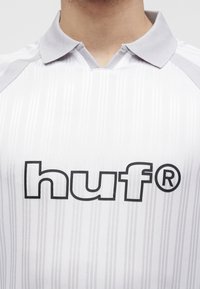 Gros plan d'un polo rayé blanc et gris avec un col gris et le logo « huf » imprimé en noir sur la poitrine.