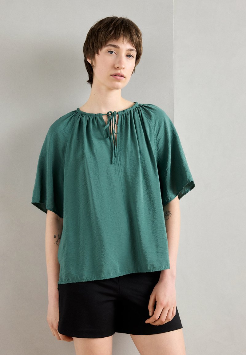 Teal short-sleeve blouse with a round neckline, gathered casing, and a tie closure in the front. Lightweight, textured fabric.