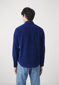 Marinblå kordflöjls skjorta med långa ärmar, tryckknappar och en spetsig yoke-design, kombinerad med ljusblå jeans, visad bakifrån.