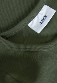 Olivgrön ribbad t-shirt krage med en vit etikett som visar "JJXX" i blått. Tygets struktur är slät, med snygga sömnadsdetaljer.