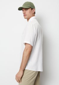 Chemise blanche à manches courtes avec une poche, en tissu texturé. Associée à une casquette verte douce, soulignant un look décontracté.