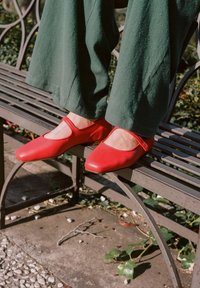 Bailarinas de piel roja con punta redondeada y corte ancho, combinadas con pantalones verdes holgados, reposando en un banco de metal al aire libre.